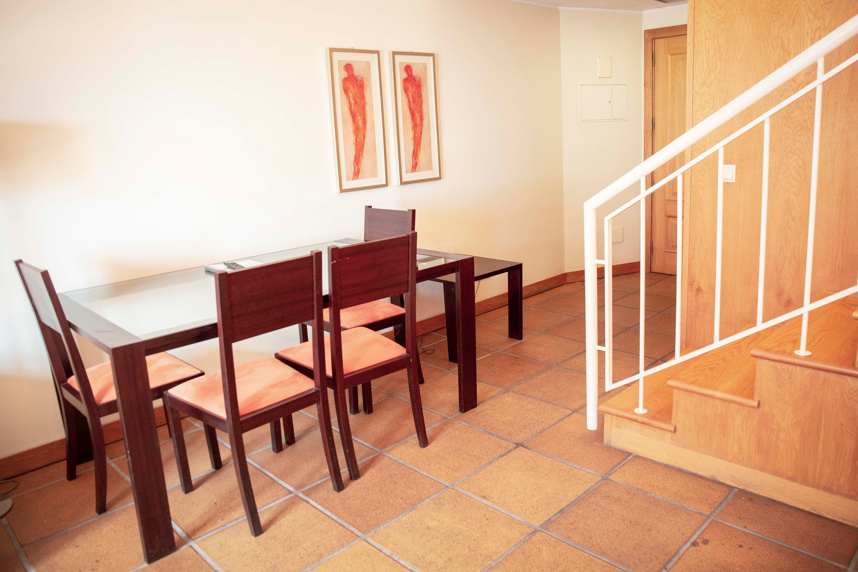 A dining area with a wooden table and chairs, a staircase with a wooden railing, and two framed artworks on the wall.