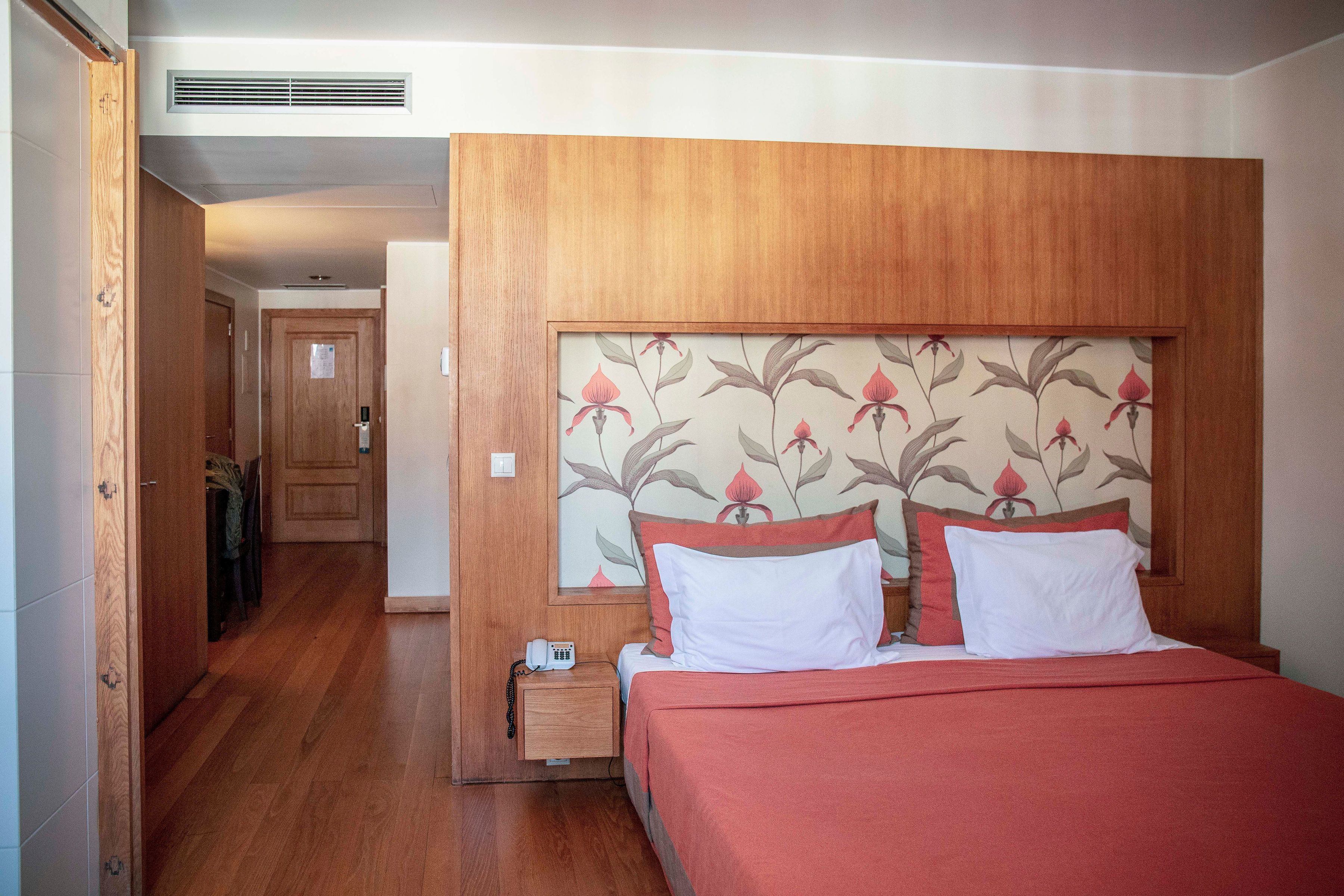 A hotel room with a wooden headboard, a bed with red bedding, a bedside table with a telephone, and a door leading to another room.