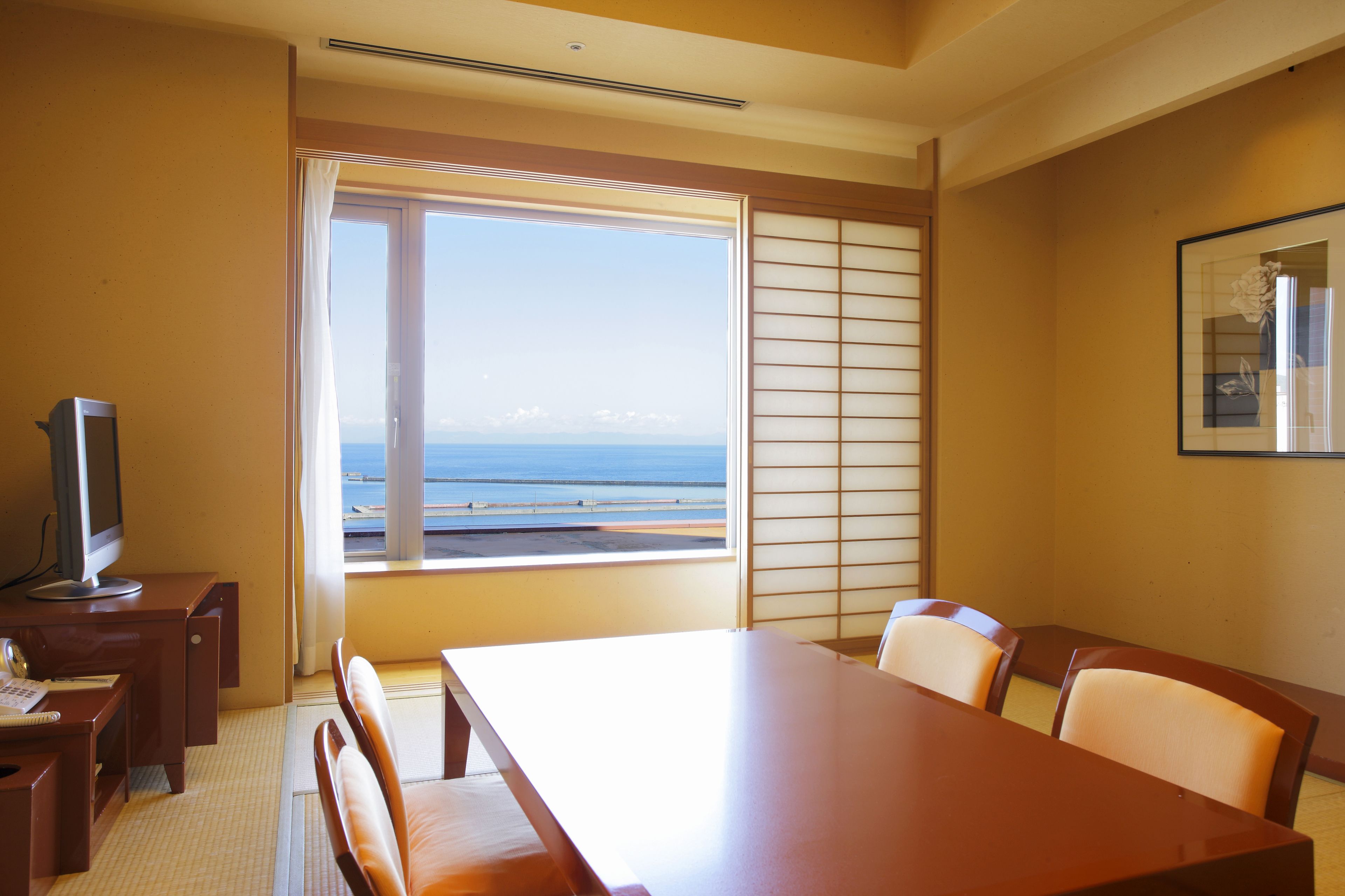 A hotel room with a large window offering a view of the sea, a wooden table, and a television on a desk.