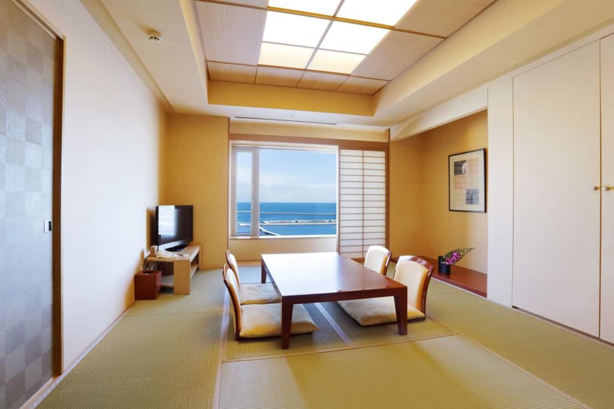 A room with tatami flooring, a low table with cushions, a television, and a large window with a view of the sea.