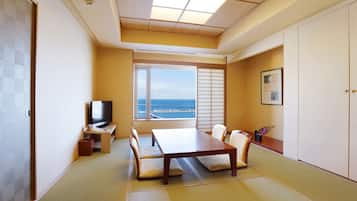 A room with tatami flooring, a low table with cushions, a television, and a large window with a view of the sea.