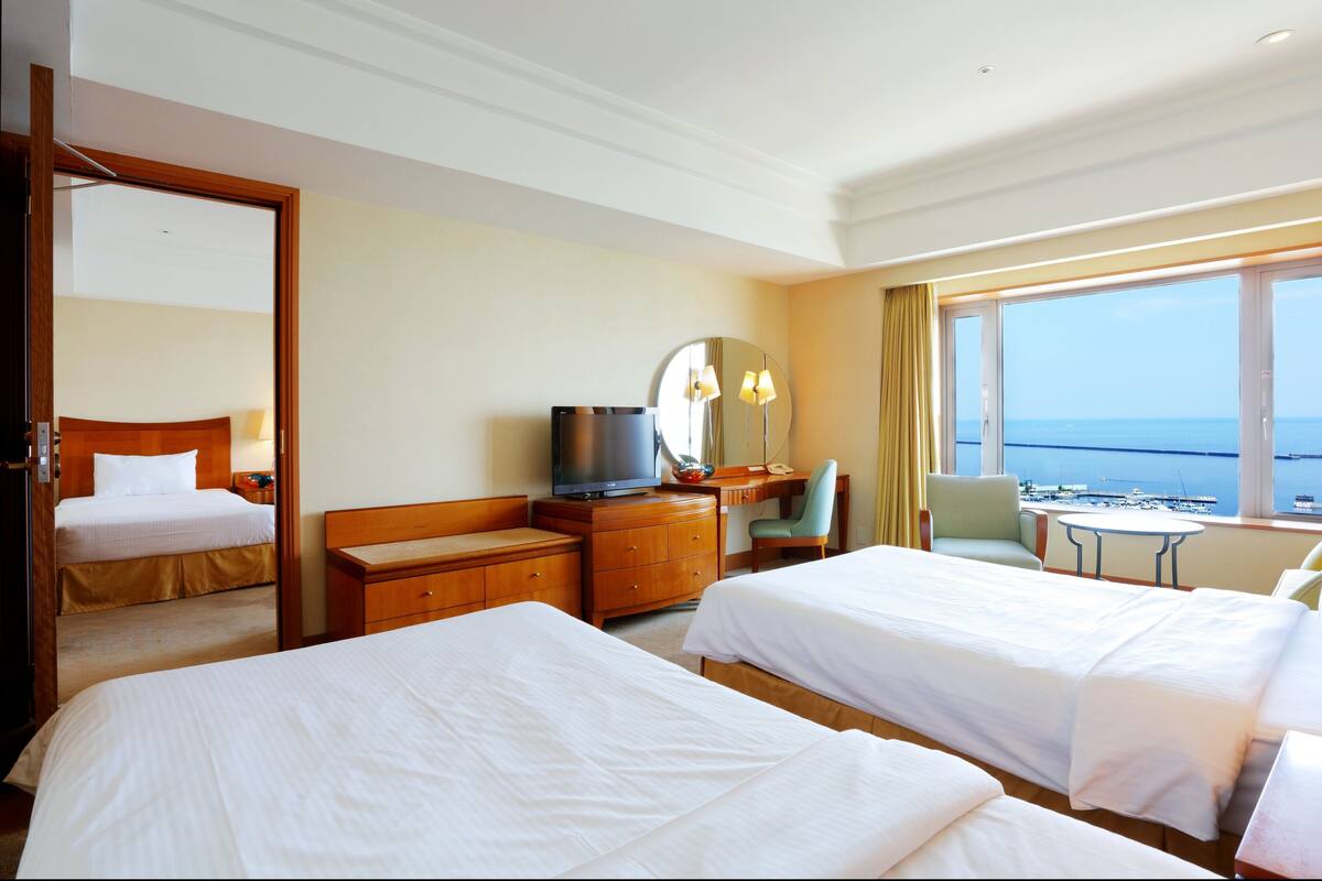 A hotel room with two beds, a desk, a chair, a television, and a view of the sea.