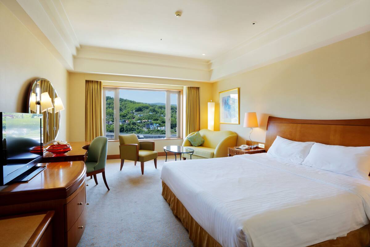 A hotel room with a large bed, a desk with a TV, a chair, a small table, and a view of a green landscape through the window.
