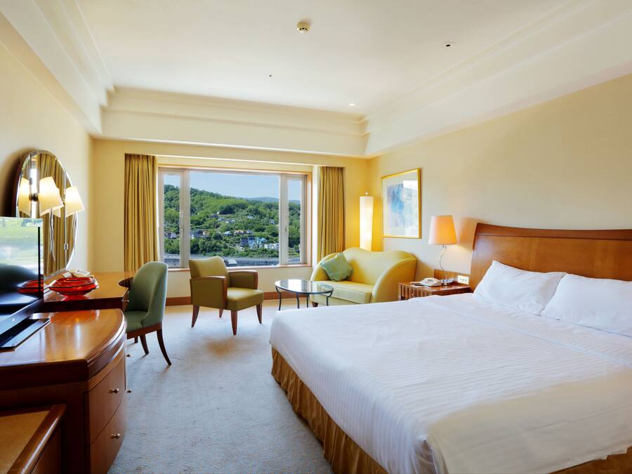 A hotel room with a large bed, a desk with a TV, a chair, a small table, and a view of a green landscape through the window.