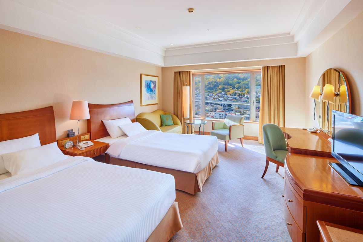 A hotel room with two beds, a desk, a chair, a television, and a view of a cityscape through large windows.