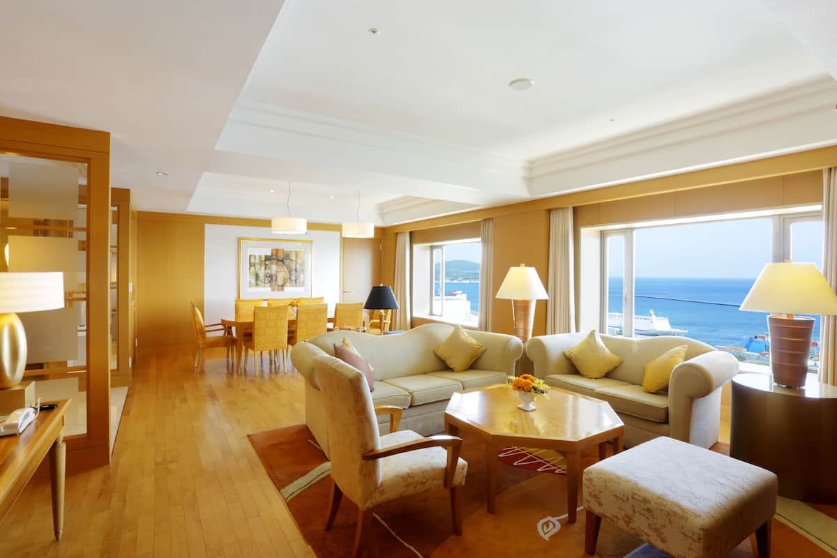 A spacious hotel room with a large window offering a view of the sea, a cozy seating area with a sofa and chairs, and a dining area with a table and chairs.