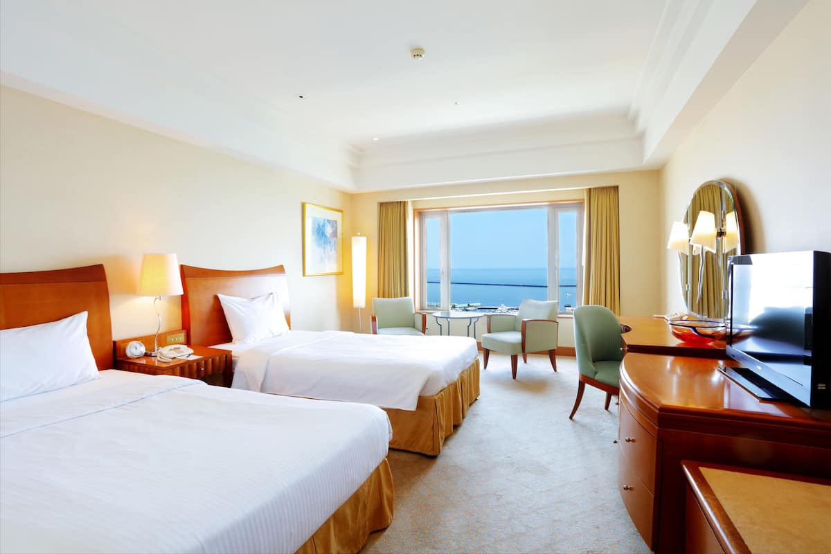 A hotel room with two beds, a desk, a chair, a television, and a view of the sea.