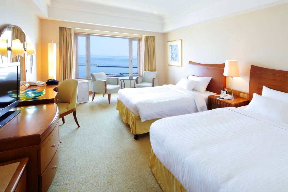 A hotel room with two beds, a desk, a chair, a table, and a view of the ocean.