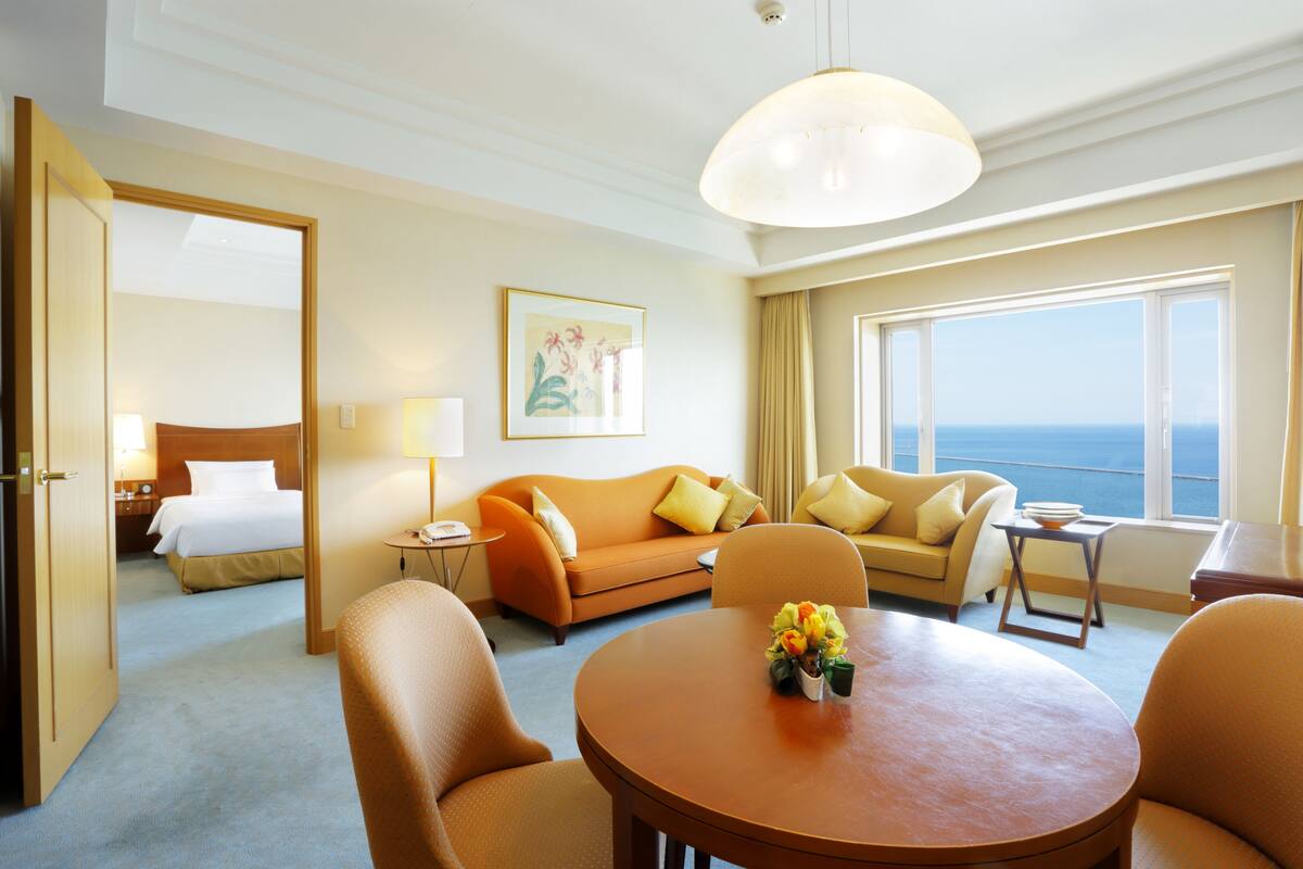 A hotel room with a bed, a sofa, a dining table, and a view of the sea.