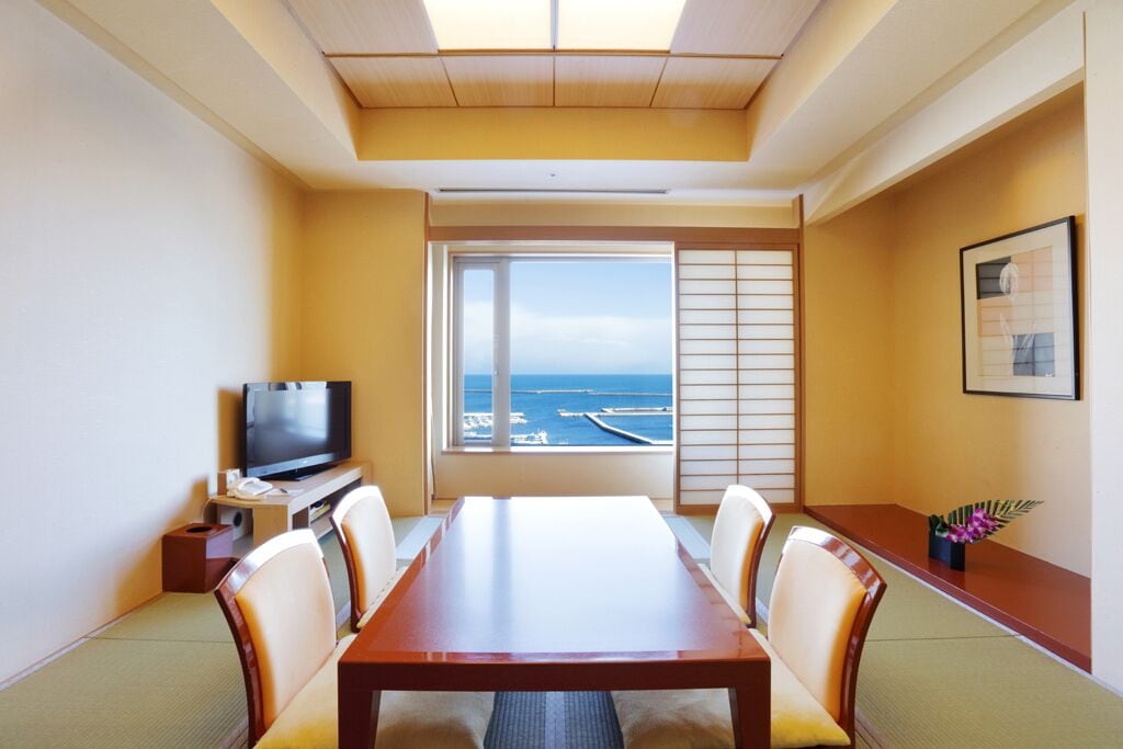 A hotel room with a wooden dining table, four chairs, a television, and a large window offering a view of the sea and boats.