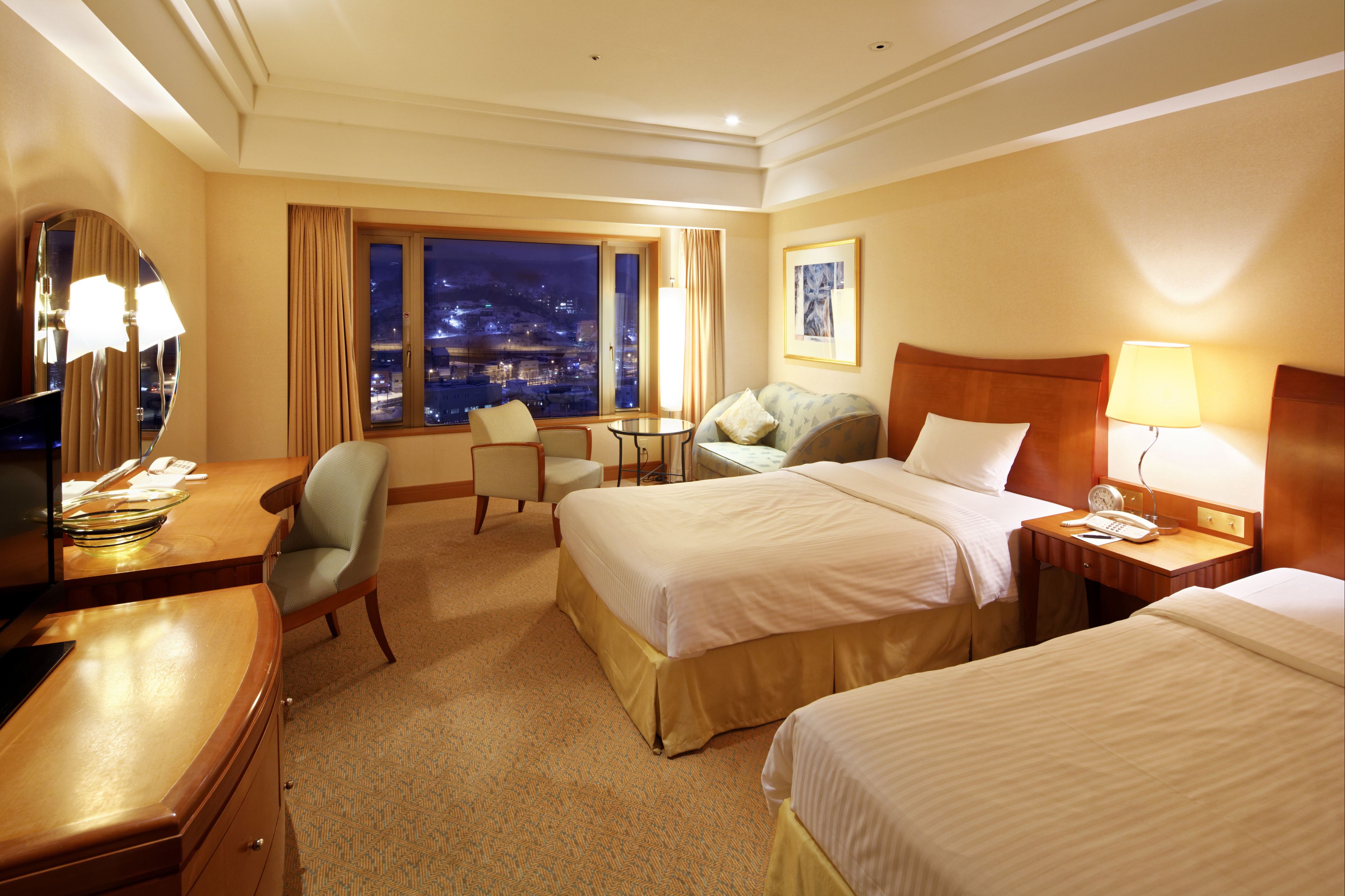 A hotel room with two beds, a desk, a chair, a small table, and a view of the city at night.