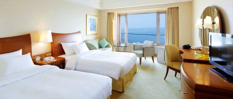 A hotel room with two beds, a desk, a chair, a TV, and a view of the sea.