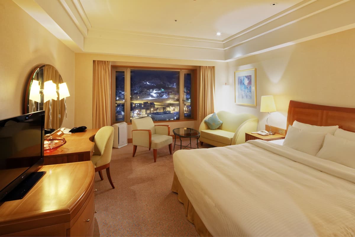A hotel room with a large bed, a desk with a computer, a chair, a small table, and a window with a night view.