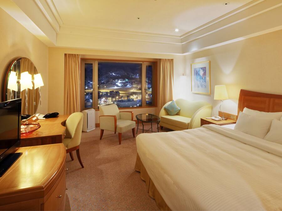 A hotel room with a large bed, a desk with a computer, a chair, a small table, and a window with a night view.