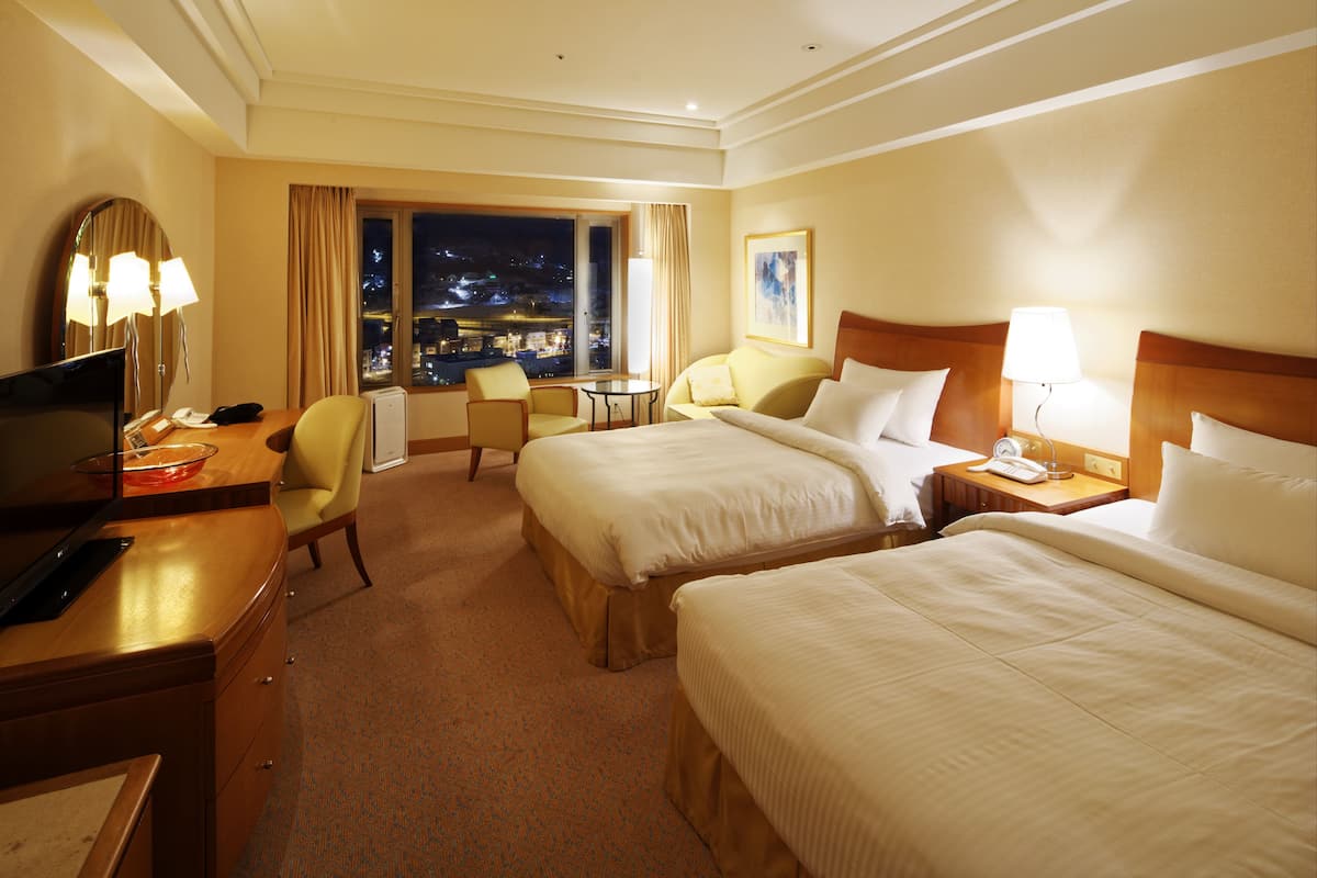 A hotel room with two beds, a desk with a computer, a chair, a small table, and a window with a city view.