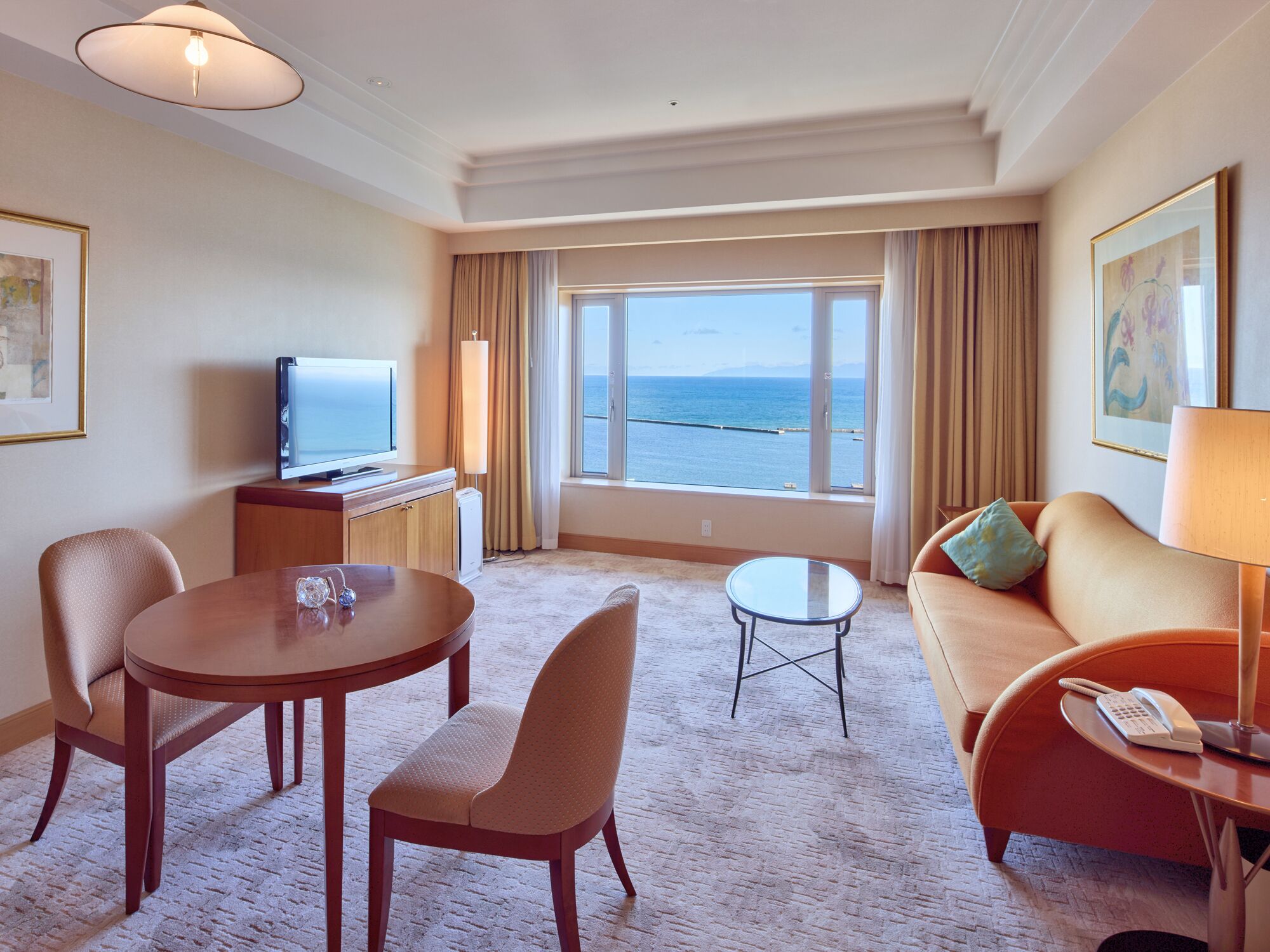 A hotel room with a large window offering a view of the sea, a sofa, a dining table with chairs, a TV, and a small side table with a lamp.