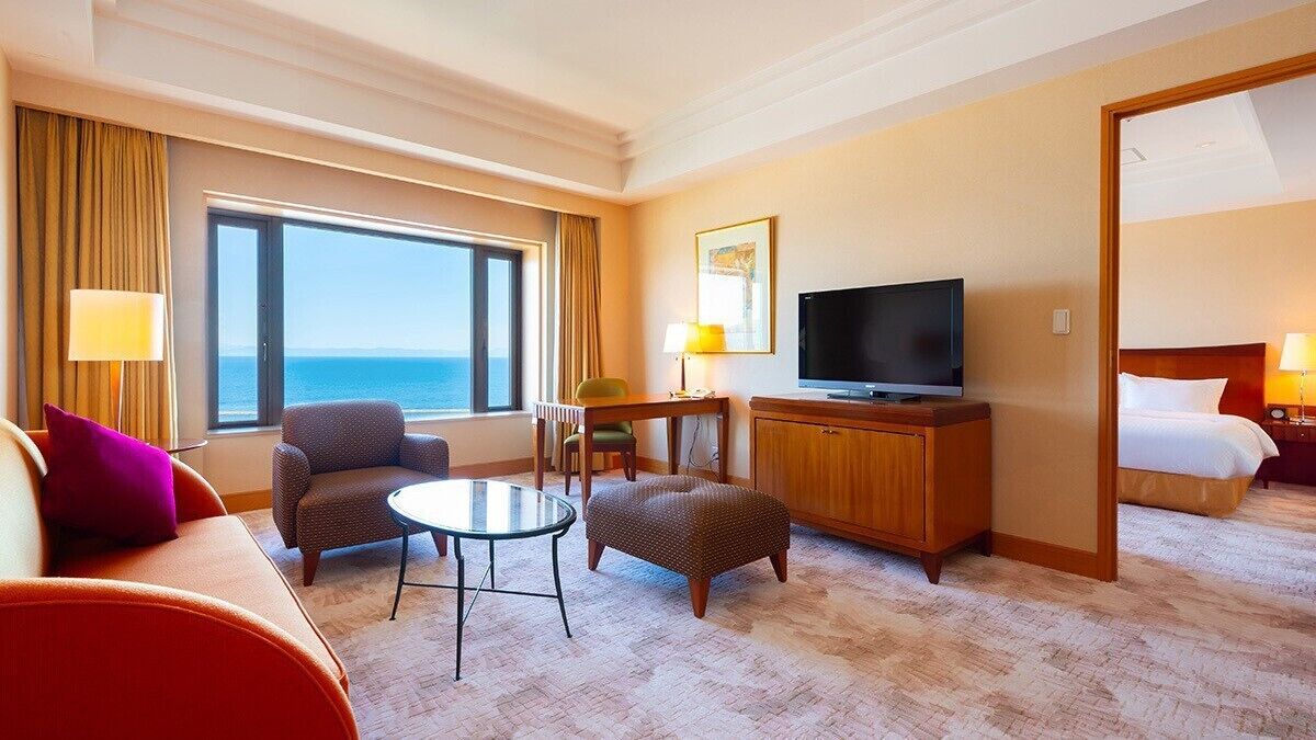 Ein Hotelzimmer mit großem Fenster, Fernseher, Sofa, Sessel, kleinem Tisch und Bett.