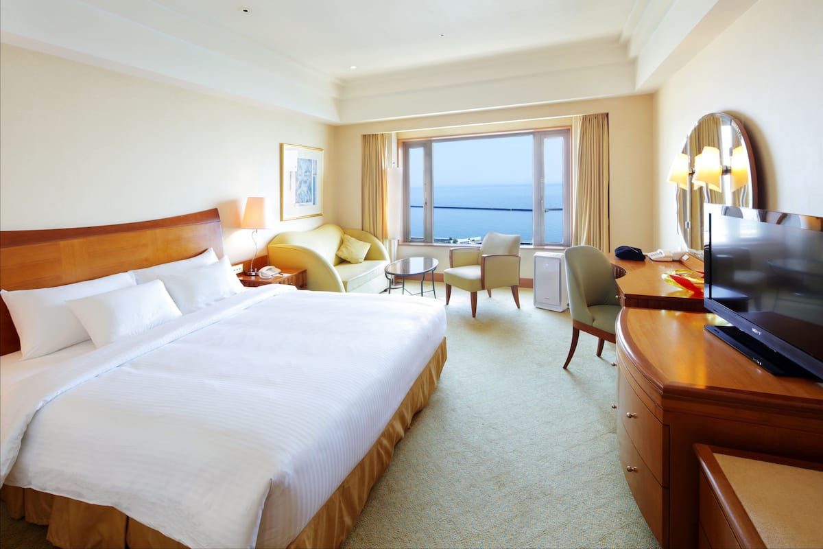 A hotel room with a large bed, a desk with a TV, a chair, a sofa, and a view of the sea.