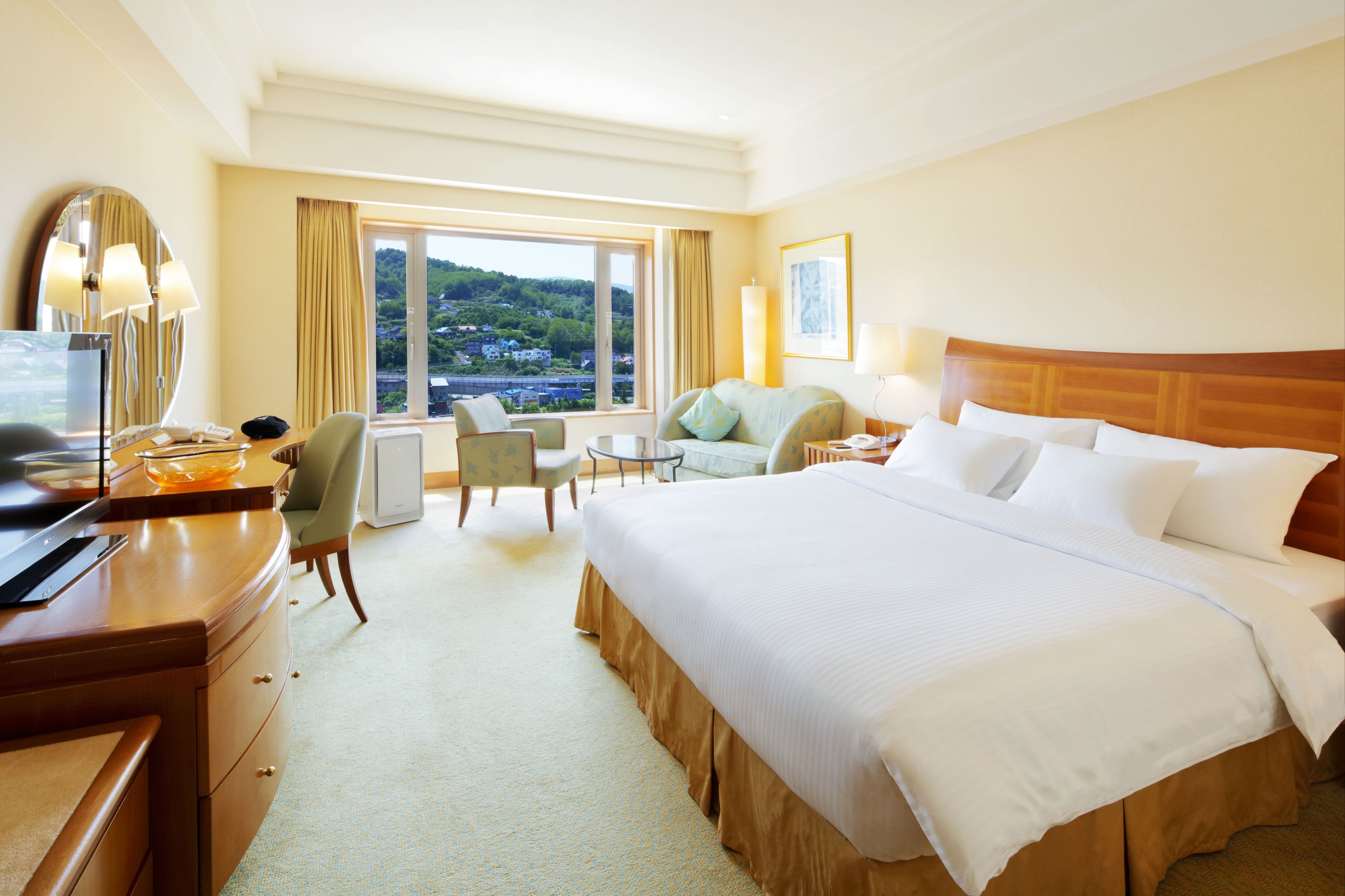A hotel room with a large bed, a desk with a computer, a chair, a sofa, and a view of a green landscape through the window.