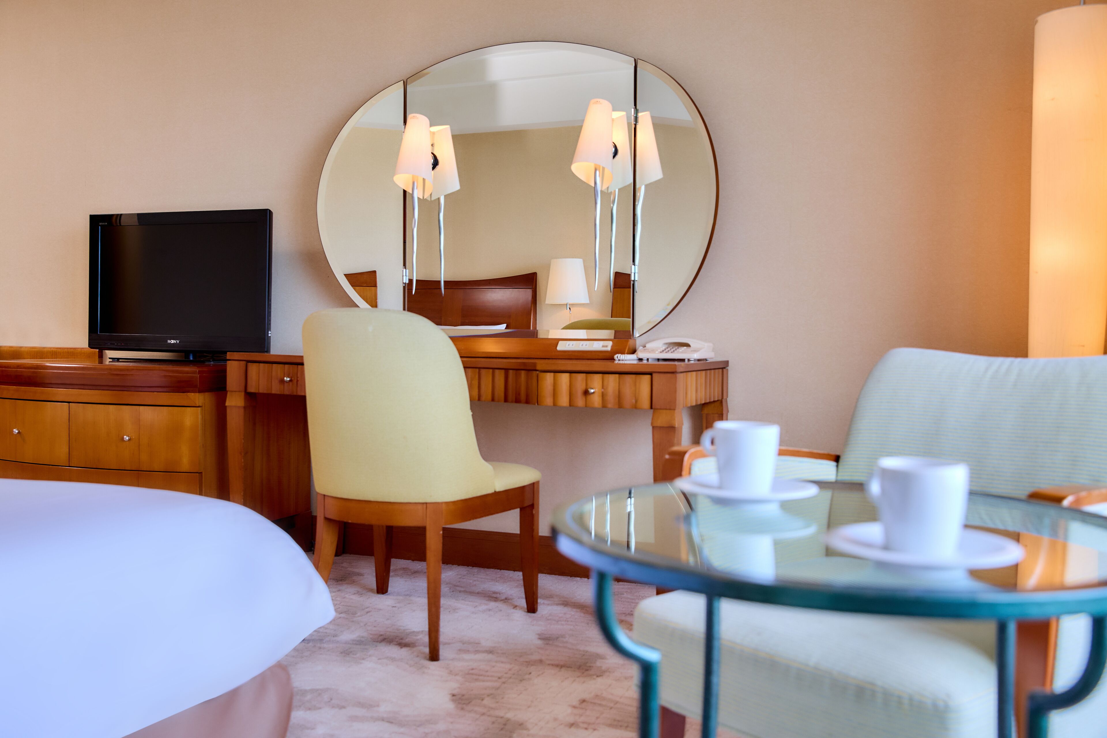 A hotel room with a bed, a desk with a computer, a chair, a round glass table with cups, and a large mirror on the wall.