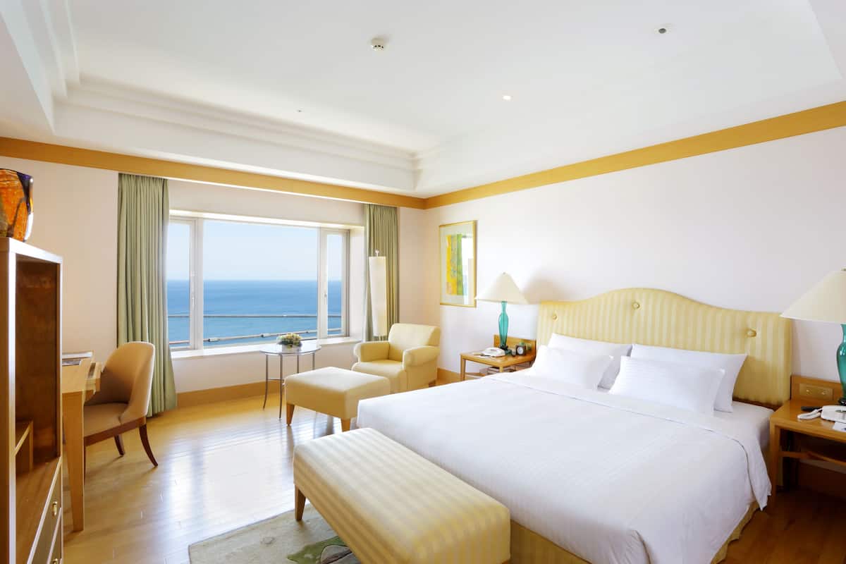 A hotel room with a large bed, a desk, a chair, a small table, and a view of the sea.