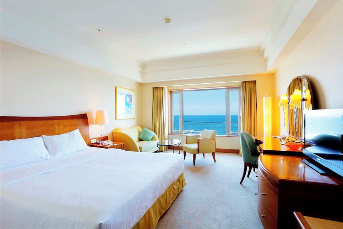 A hotel room with a large bed, a desk, a chair, a small table, and a view of the ocean.