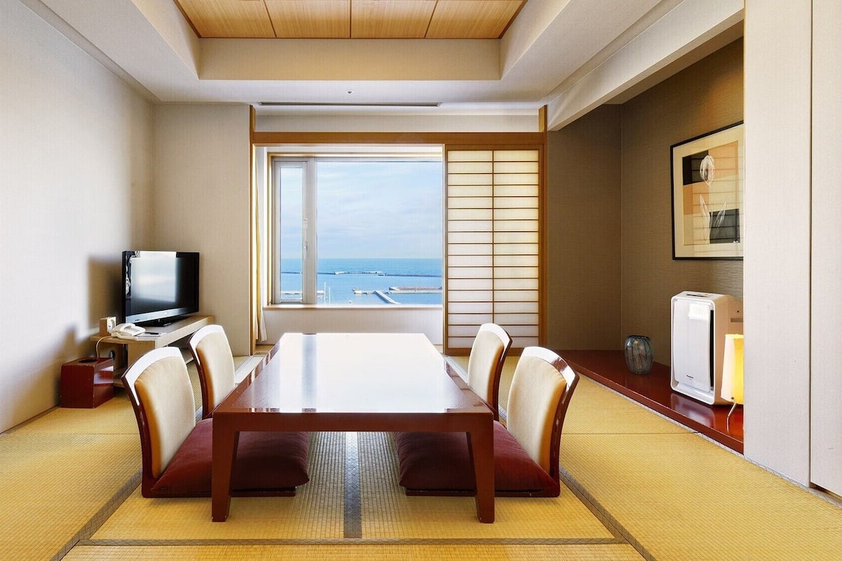 Habitación de hotel con un ventanal grande, una mesa de comedor con sillas, un televisor y una pequeña kitchenette.