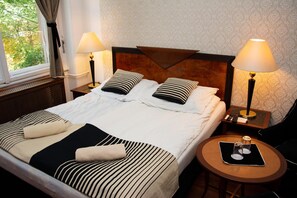 Een net opgemaakt bed met gestreept beddengoed, twee nachtlampen, een klein rond tafeltje met een glas en uitzicht op groen buiten.