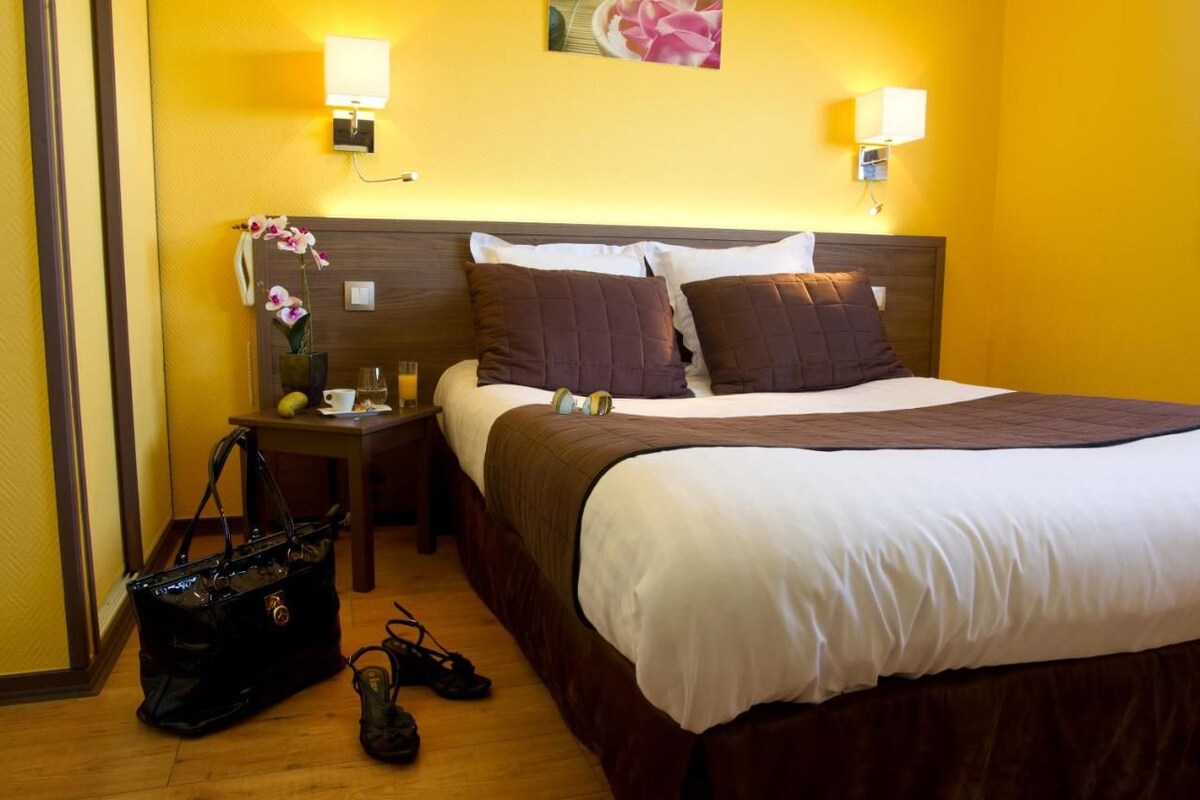 Una habitación de hotel con una cama, mesitas de noche, una lámpara, un bolso de mano y un par de zapatos.
