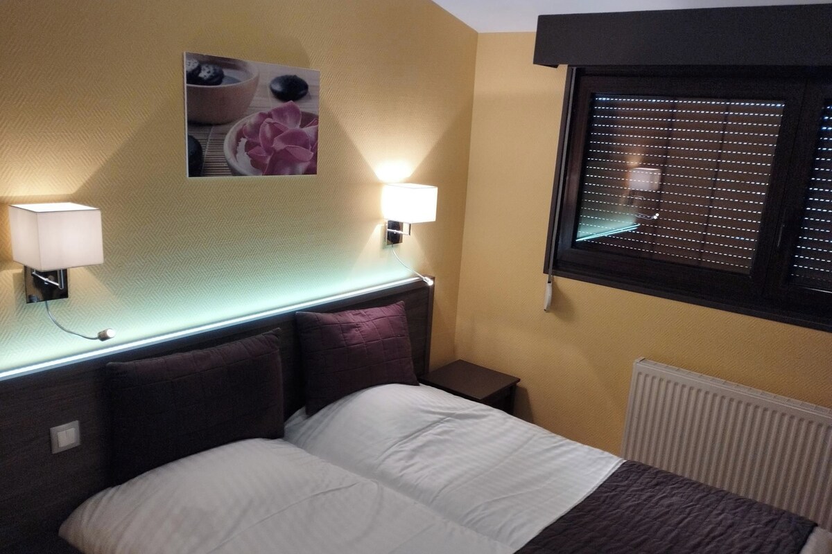 Una habitación de hotel con cama, dos lámparas de noche, un cuadro en la pared, una ventana con persianas y un radiador.