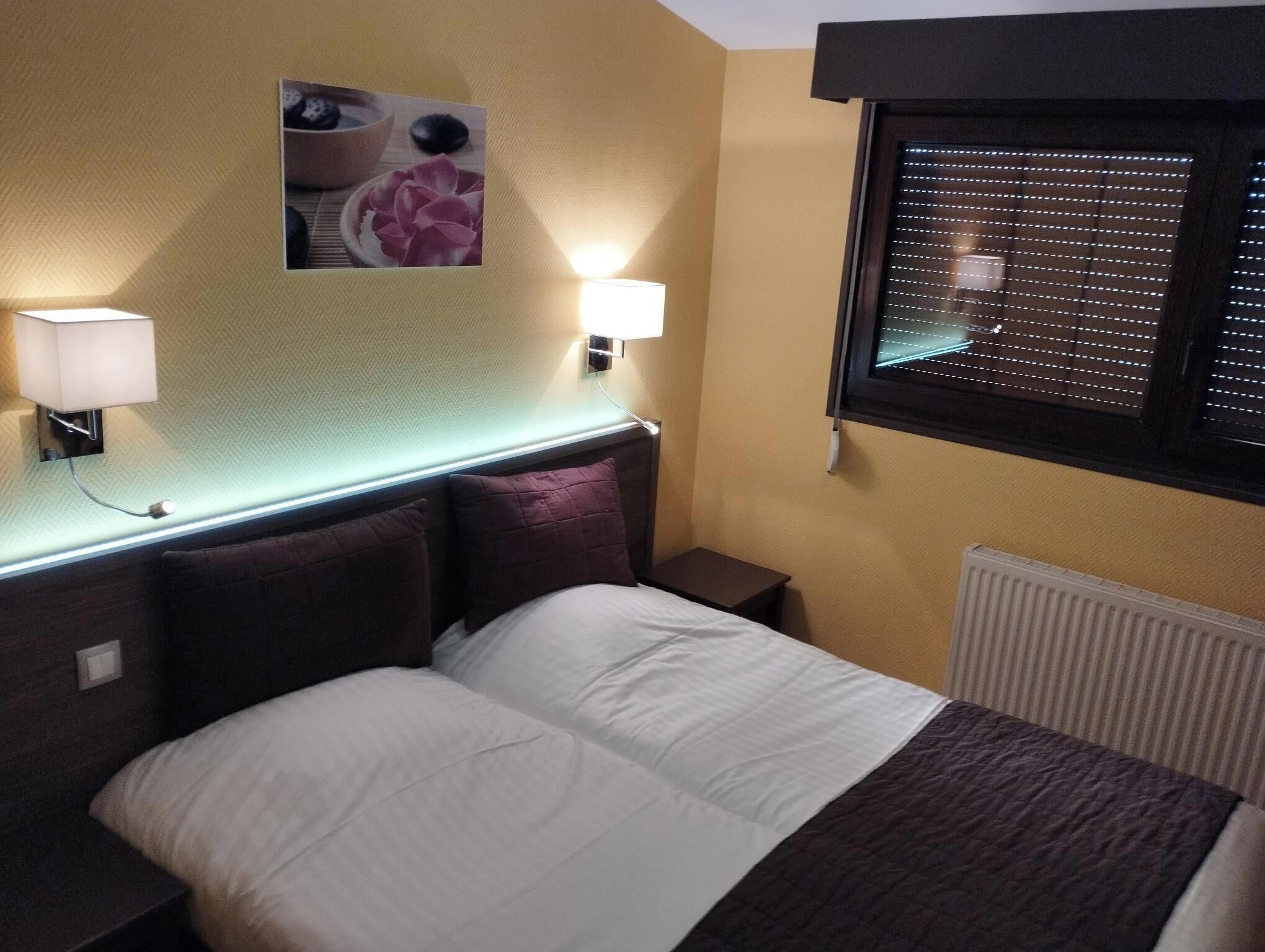 Une chambre d’hôtel avec un lit, deux lampes de chevet, un tableau accroché au mur, une fenêtre avec des stores et un radiateur.