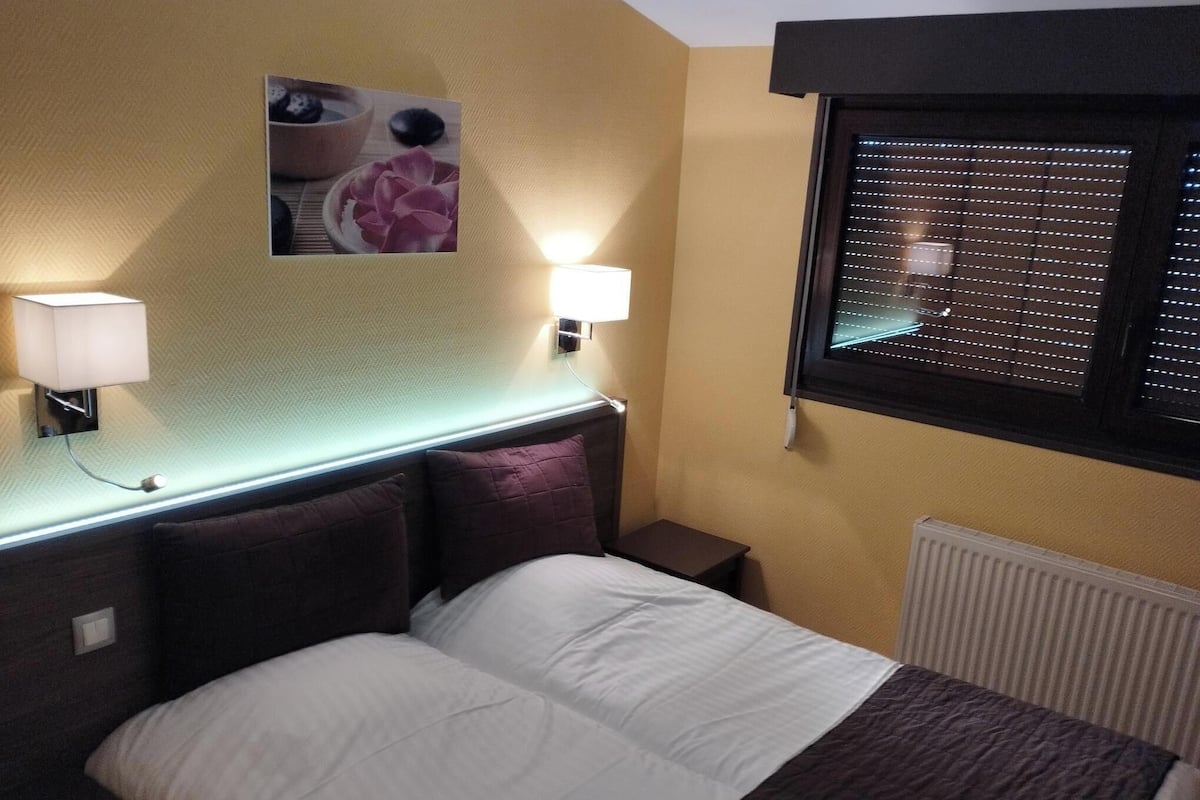 Una habitación de hotel con cama, dos lámparas de noche, un cuadro en la pared, una ventana con persianas y un radiador.
