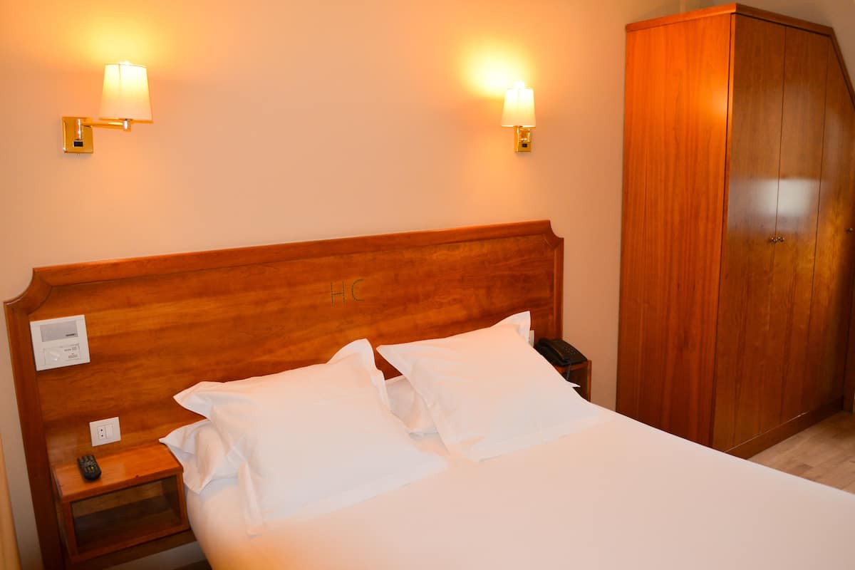 Quarto de hotel com cabeceira de madeira, duas luminárias laterais, telefone ao lado da cama e um guarda-roupa.