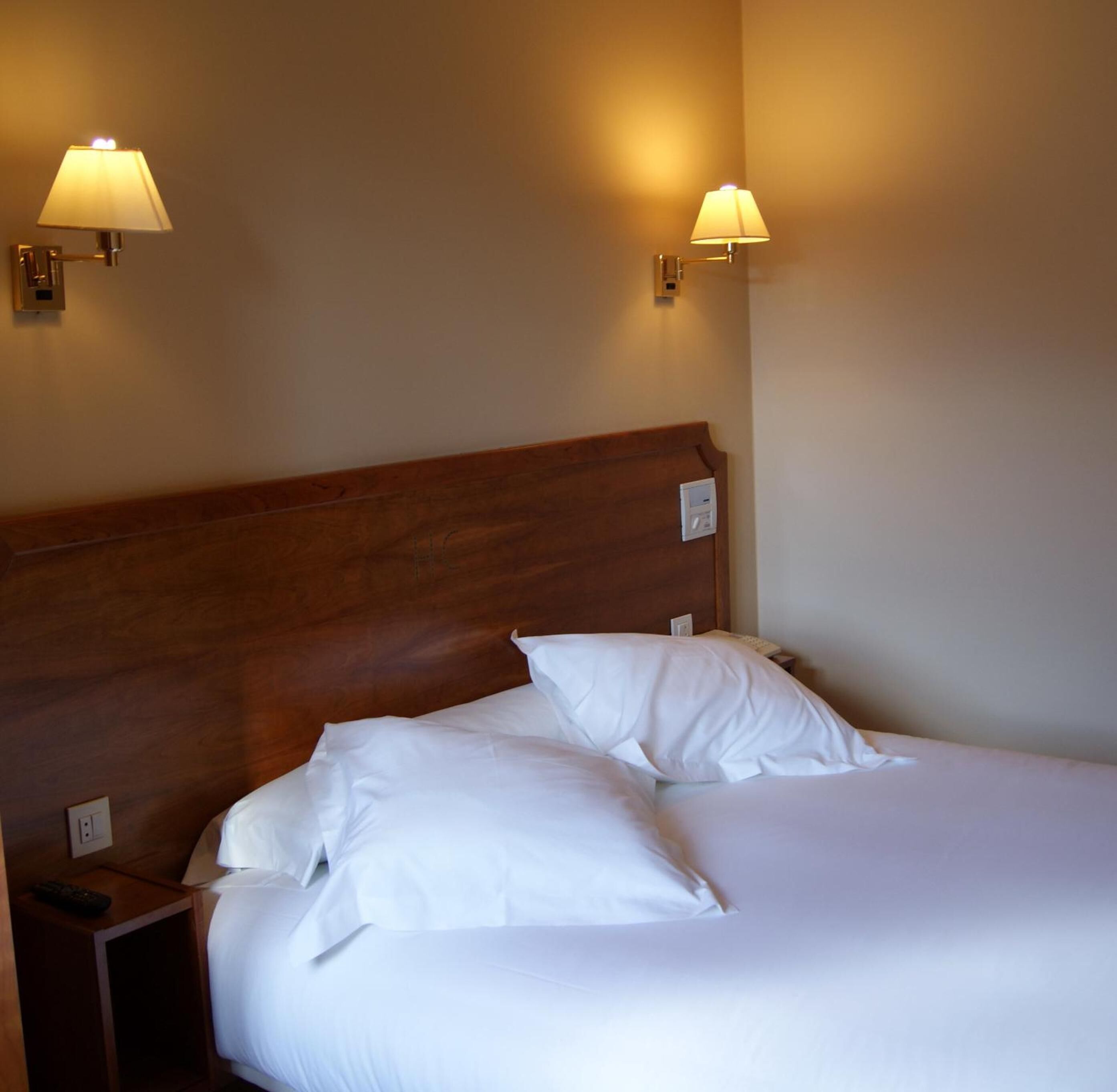 Una cama bien hecha con sábanas blancas y dos almohadas, un cabecero de madera y dos lámparas de pared a cada lado.