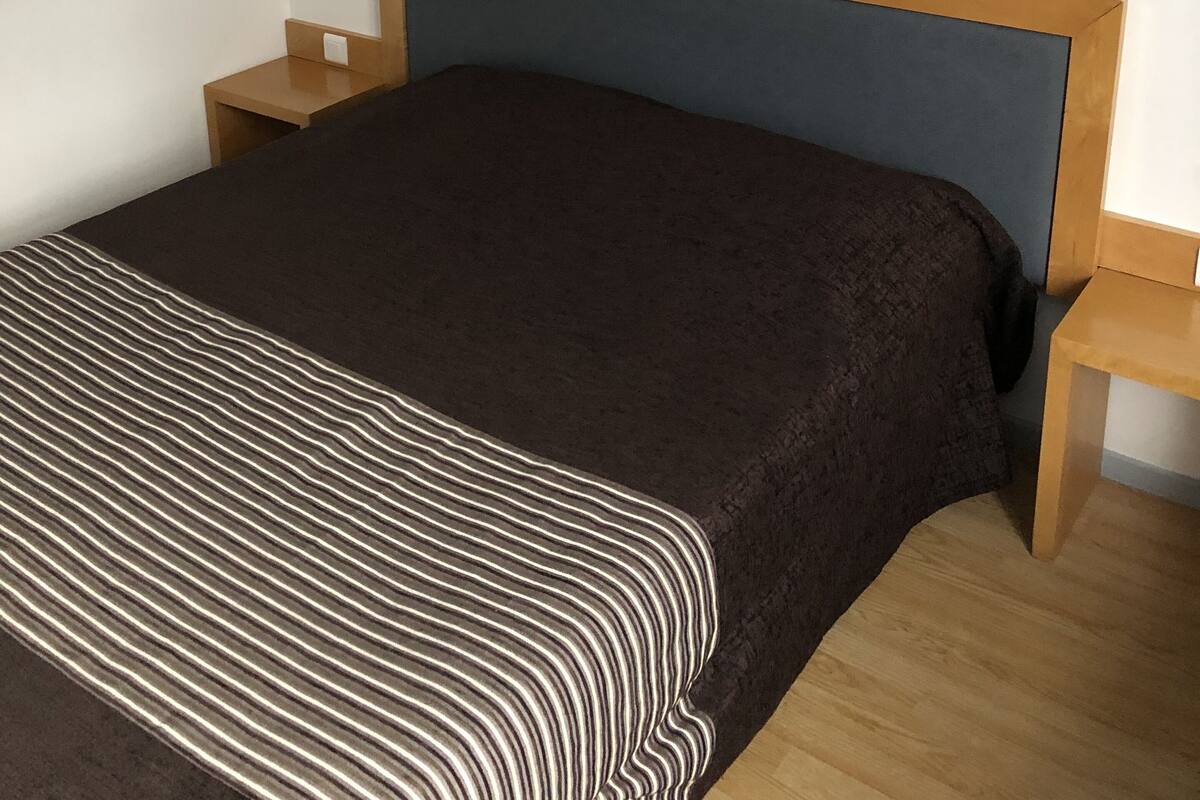 Una cama bien hecha con una colcha a rayas, dos mesitas de noche con lámparas y un cuadro en la pared.