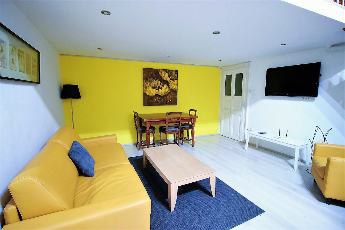 A living room with a yellow sofa, a wooden coffee table, a dining table with chairs, a painting on the wall, and a TV mounted on the wall.