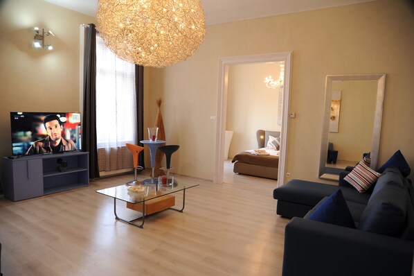 A modern living room with a flat-screen TV, a glass coffee table, a dark sofa, and a large decorative light fixture. - Budapest Easy Flats- Oktogon Lux Apartment (Budapest)