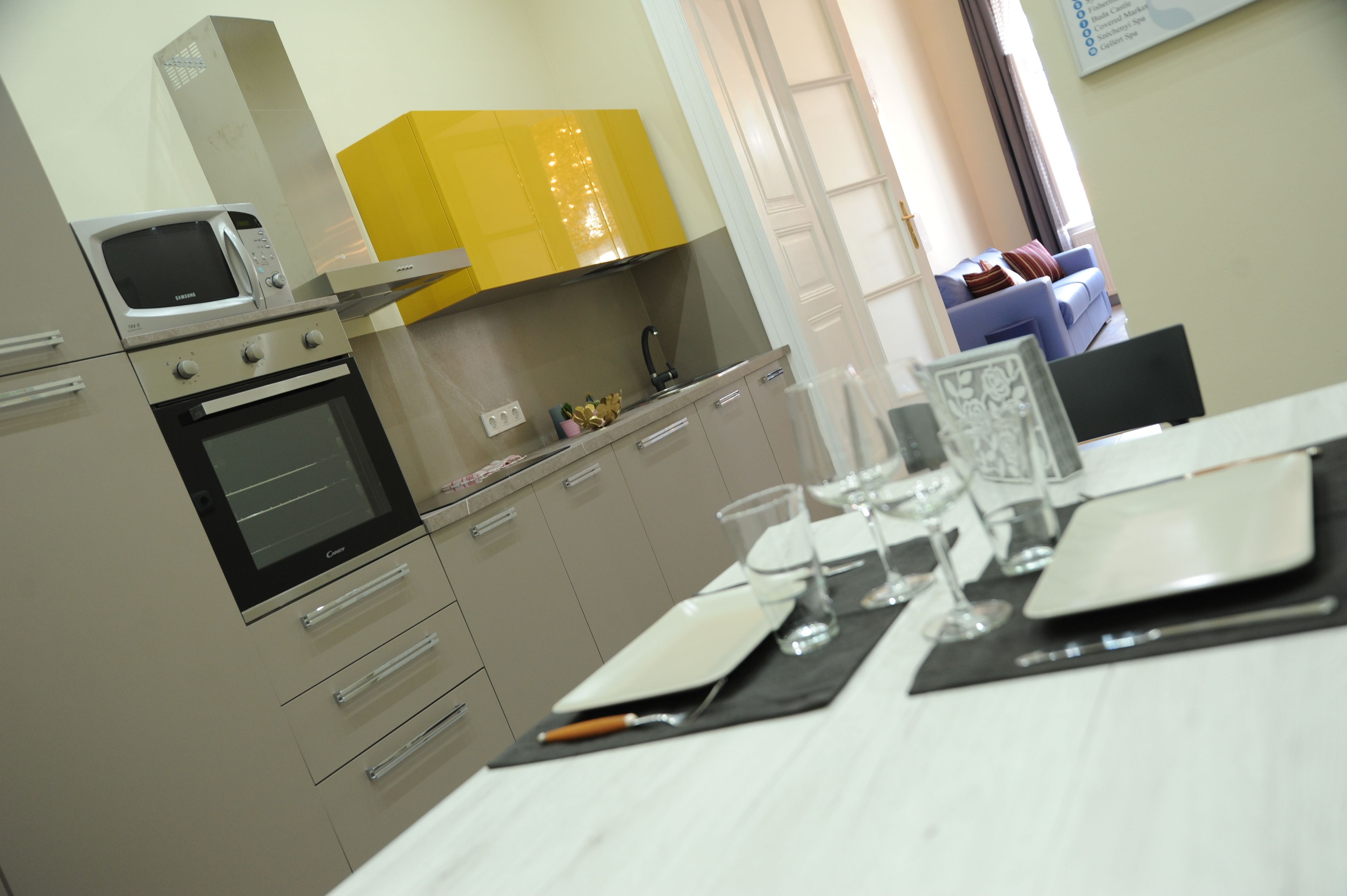 A modern kitchen with a microwave, oven, and yellow cabinets. There is a dining table set with plates, glasses, and cutlery.