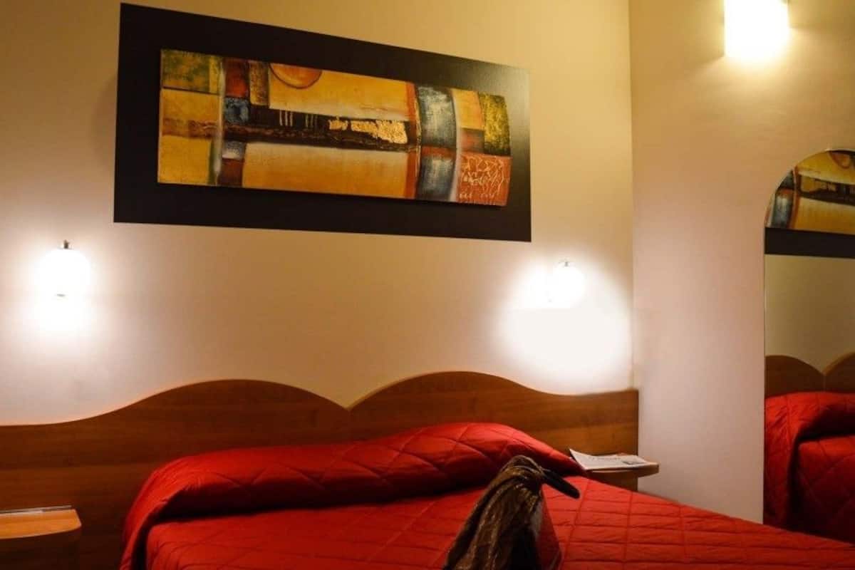 A hotel room with a wooden headboard, a red bedspread, a painting above the bed, and two wall-mounted lights.