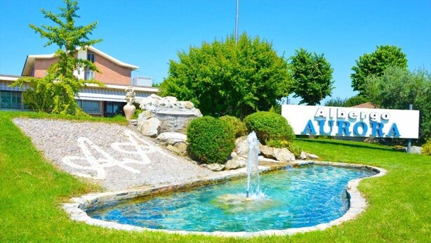 A swimming pool with a fountain, surrounded by a well-maintained lawn and a building with a sign reading "Albergio Aurora". - Albergo Aurora (Castenaso)