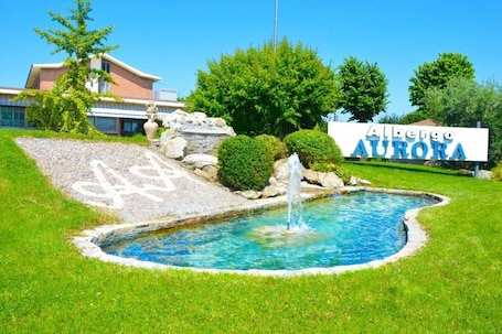 A swimming pool with a fountain, surrounded by a well-maintained lawn and a building with a sign reading "Albergio Aurora".