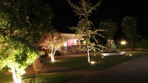 A well-lit garden area with a fountain, a large planter, and a building with steps leading up to a door. - Albergo Aurora (Castenaso)