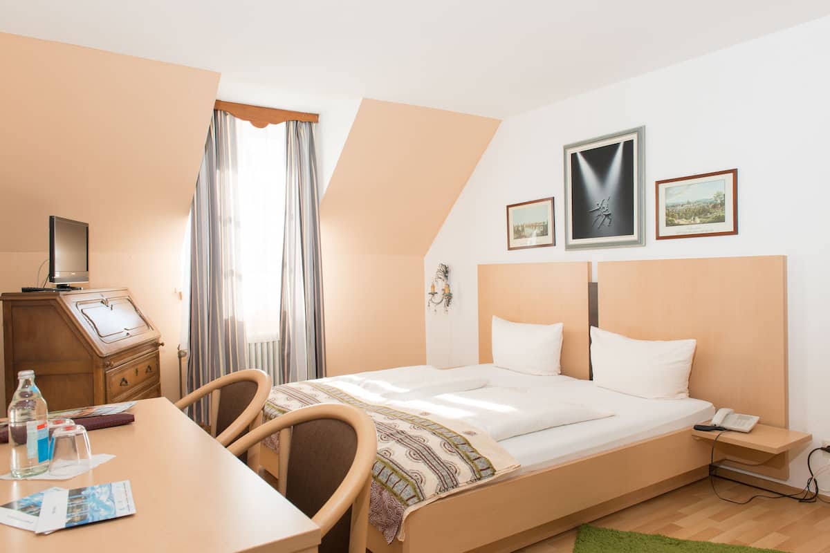 Ein Hotelzimmer mit Bett, Schreibtisch, Stuhl und einem kleinen Tisch mit Flasche und Unterlagen.