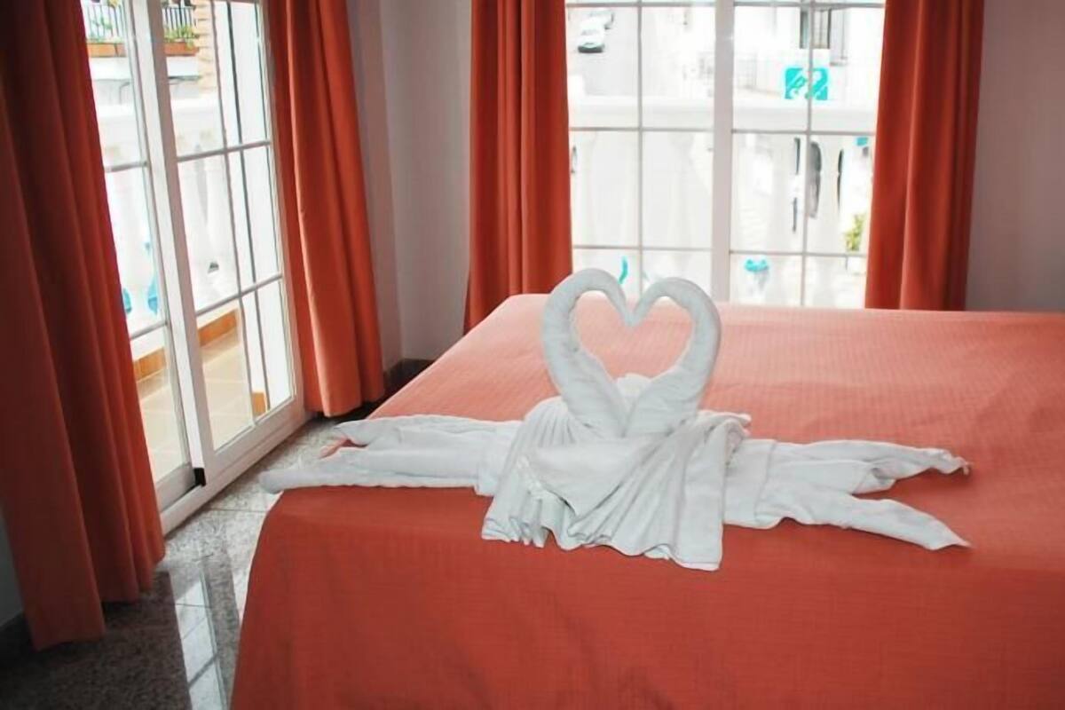 A hotel room with a bed, orange curtains, and a window with a view of the outside.