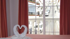 A hotel room with a bed, a heart-shaped towel arrangement, and a view of a street with parked cars and buildings. - Mary Tere (Salobreña)