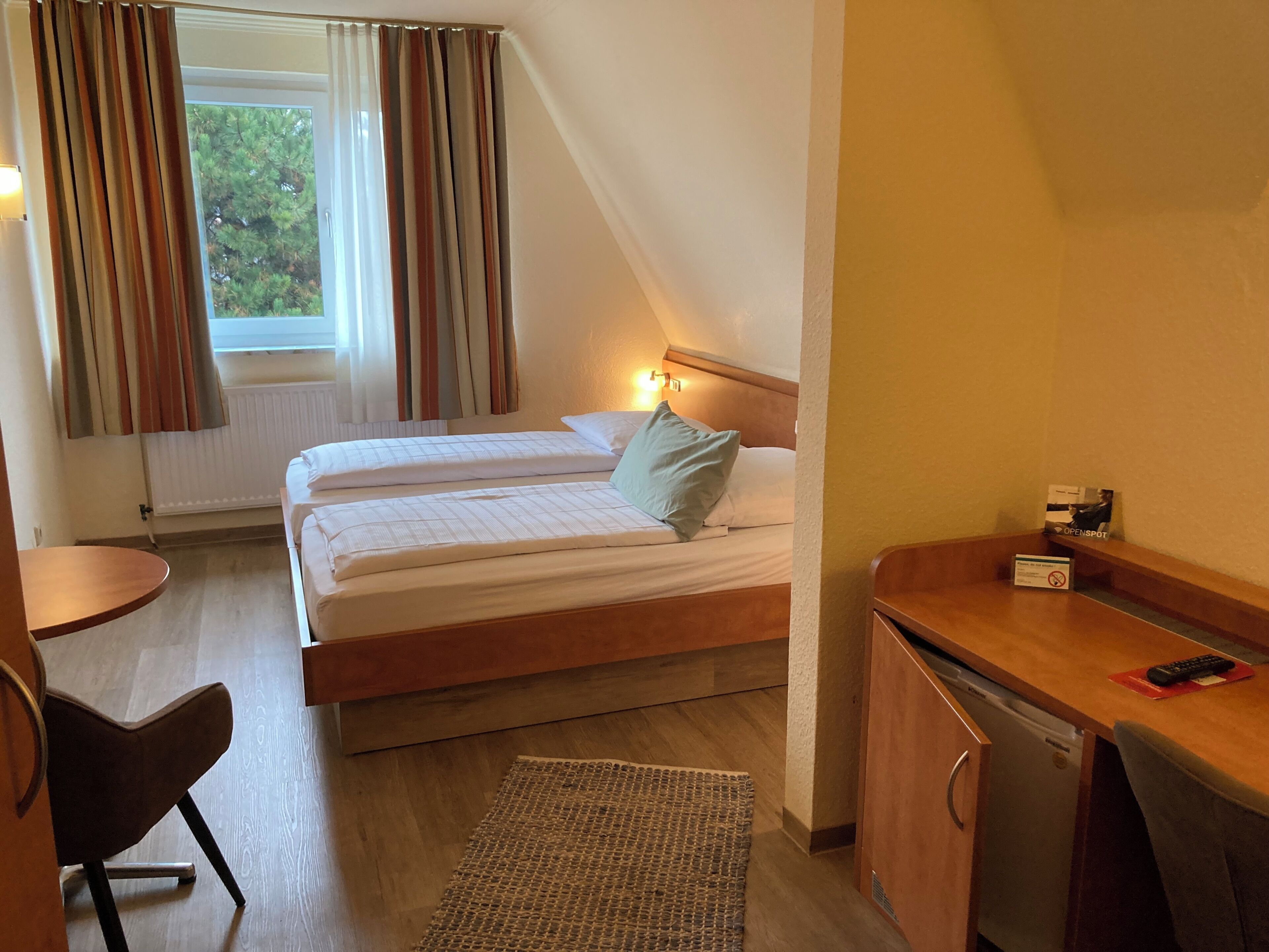 Een hotelkamer met twee bedden, een bureau, een stoel, een kleedje en uitzicht op bomen door het raam.