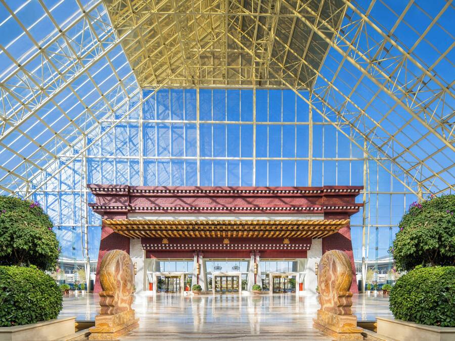 A spacious atrium with a glass ceiling, a central entrance with a red and gold structure, and symmetrical landscaping with large stone sculptures.