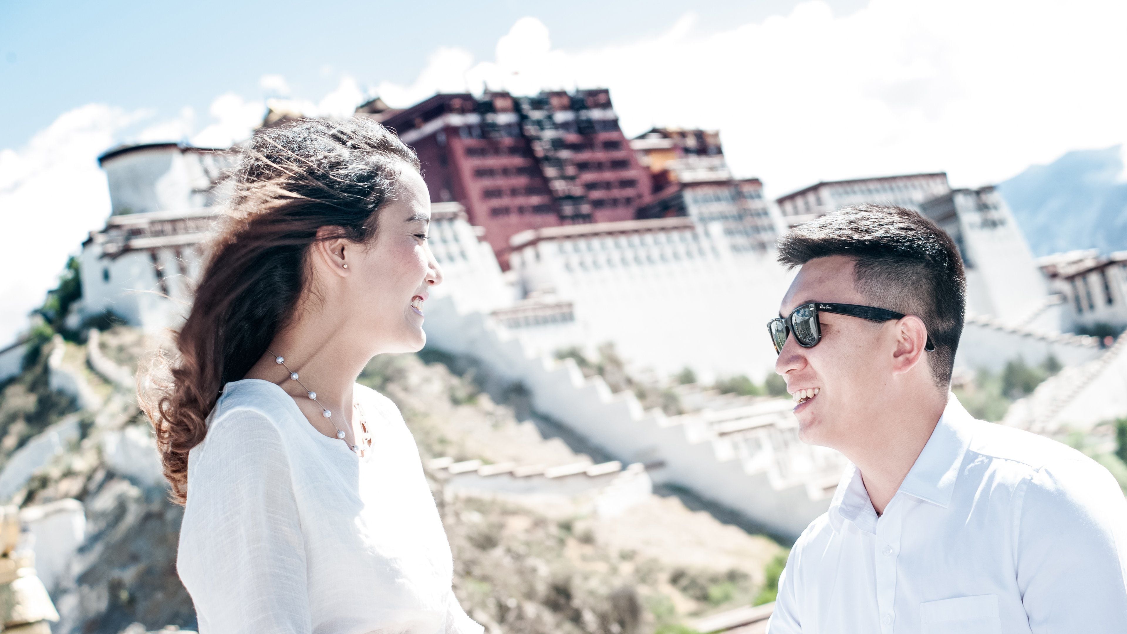 Un couple se tient devant un bâtiment historique à l’architecture traditionnelle, avec une imposante structure en forme de forteresse en arrière-plan.