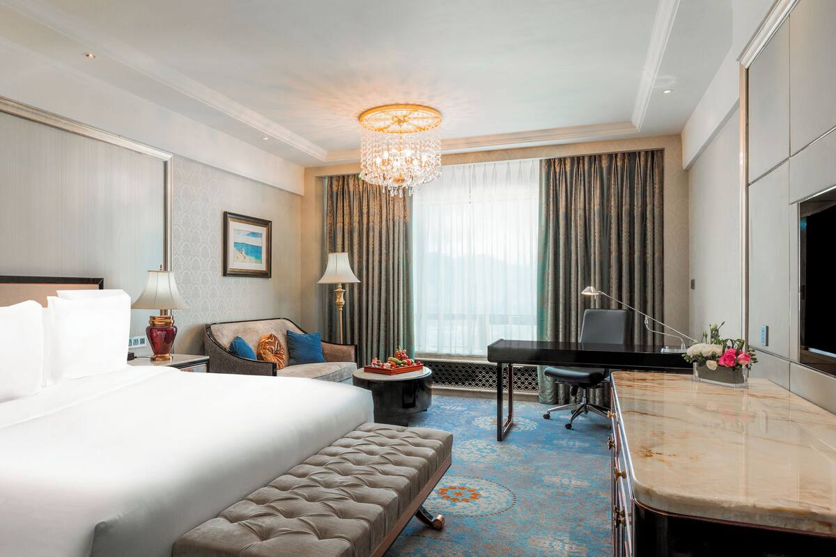 Quarto de hotel com cama grande, escrivaninha, cadeira, luminária de mesa, televisão e janela com cortinas.