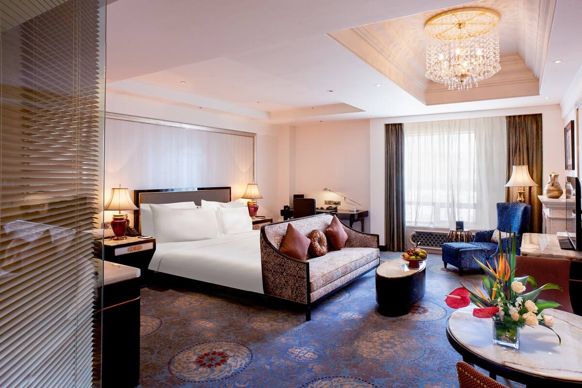Quarto de hotel com cama, sofá, escrivaninha, cadeira, mesa de jantar e lustre.